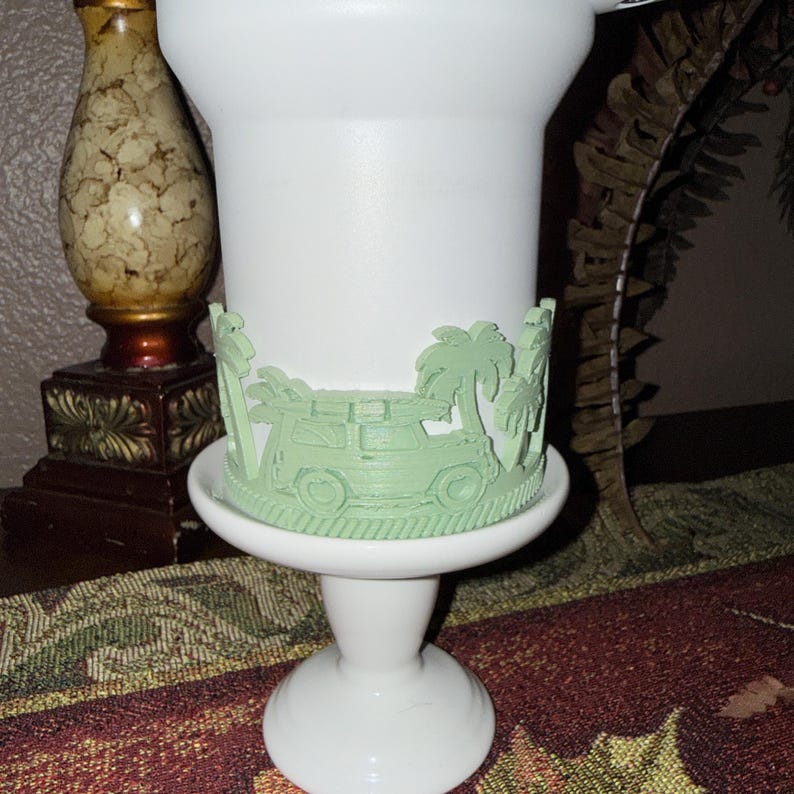 May include: A white pedestal cup featuring a light green wrap-around relief design. The raised decoration depicts a vehicle with a surfboard on its roof, surrounded by palm trees. The cup has a smooth finish and a flared base, standing on a stem.