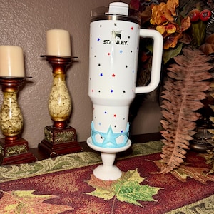 May include: White Stanley tumbler with a handle, decorated with red, blue, and black stars. The tumbler has the Stanley logo and is placed on a white pedestal with a light blue star-shaped design.