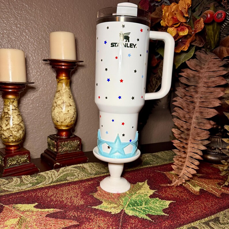 May include: White Stanley tumbler with a handle, decorated with red, blue, and black stars. The tumbler has the Stanley logo and is placed on a white pedestal with a light blue star-shaped design.