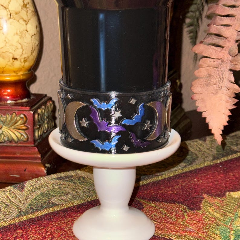 May include: A black candle holder with a decorative band featuring silver moons, purple and blue bats, and stars. The candle holder sits on a white pedestal base, creating a spooky, Halloween-themed aesthetic.