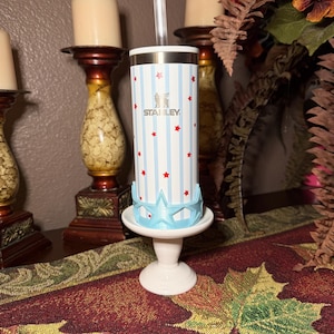 May include: A Stanley tumbler with a white, blue, and red star and stripe design. The tumbler is on a white pedestal with a blue star-shaped base. The tumbler has a clear straw and the Stanley logo.