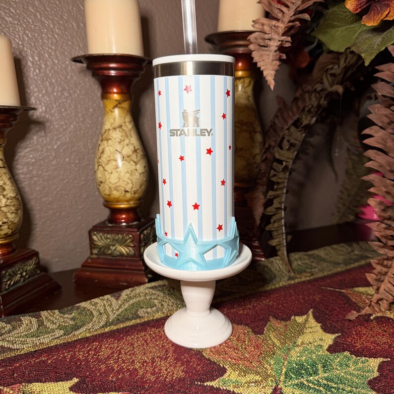 May include: A Stanley tumbler with a white, blue, and red star and stripe design. The tumbler is on a white pedestal with a blue star-shaped base. The tumbler has a clear straw and the Stanley logo.