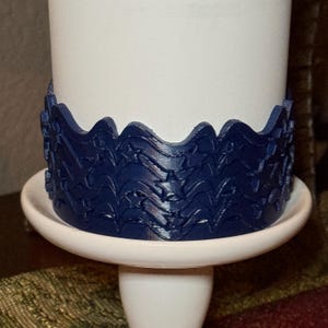 May include: A dark blue, circular, decorative band featuring a repeating cutout pattern of stars and wavy lines. The band is wrapped around the base of a smooth, white cylindrical object that sits on a white, pedestal-style circular base.