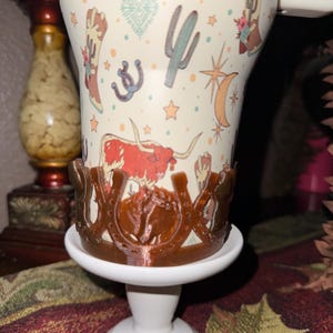 K&ouml;nnte beinhalten: Eine wei&szlig;e Keramiktasse mit Cowboy-Motiv, die einen Longhorn, einen Kaktus und ein Hufeisen zeigt. Die Tasse wird von einem braunen hufeisenf&ouml;rmigen Halter auf einem wei&szlig;en Sockel gehalten. Die Tasse hat einen Henkel und ist mit Sternen verziert.