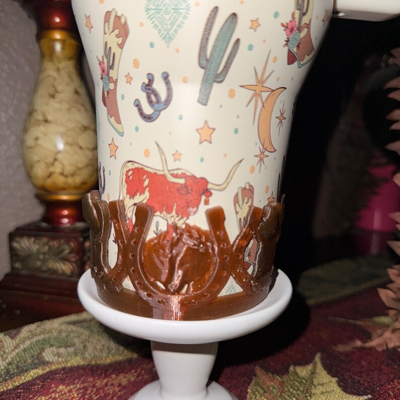 K&ouml;nnte beinhalten: Eine wei&szlig;e Keramiktasse mit Cowboy-Motiv, die einen Longhorn, einen Kaktus und ein Hufeisen zeigt. Die Tasse wird von einem braunen hufeisenf&ouml;rmigen Halter auf einem wei&szlig;en Sockel gehalten. Die Tasse hat einen Henkel und ist mit Sternen verziert.
