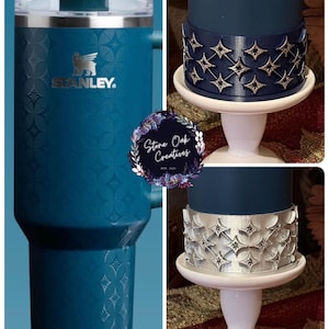May include: A teal Stanley tumbler with a silver rim and handle, featuring the Stanley logo. The tumbler has a repeating geometric pattern. Also shown are decorative bands with starburst designs, in silver and navy blue, displayed on white pedestals.