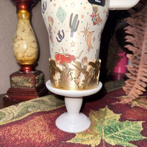 K&ouml;nnte beinhalten: Ein wei&szlig;er Becher mit Western-Motiv mit Kakteen, Stiefeln und einem Longhorn. Der Becher steht auf einem wei&szlig;en Sockel mit einem goldfarbenen, dekorativen Rand. Der Becher hat etwa 1,1 Liter.
