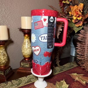 May include: A 40oz stainless steel tumbler with a denim-textured blue wrap featuring stickers of an American flag, a white daisy, a heart with the text DARLIN, cherries, and dice. It has a red lid, a red handle, and a red base accent.