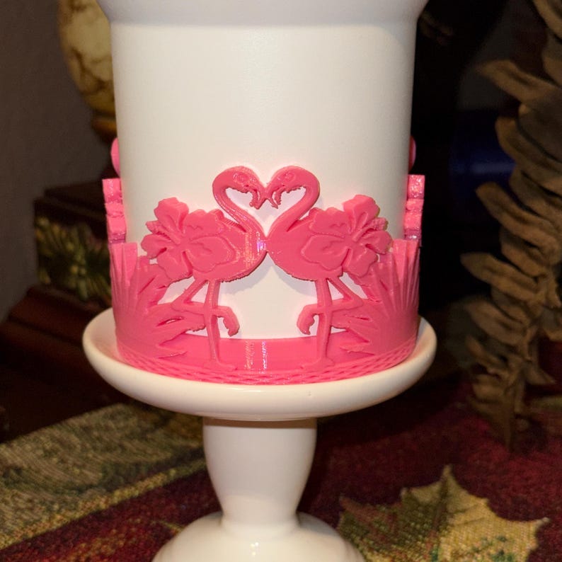 May include: A white cylindrical candle holder featuring a raised, bright pink decorative wrap. The wrap displays two flamingos facing each other with their necks forming a heart shape, surrounded by hibiscus flowers and tropical foliage patterns.