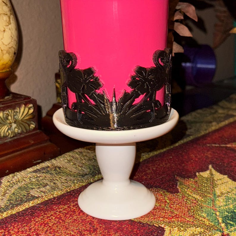 May include: A bright pink cylindrical candle sits inside a black decorative sleeve featuring a cutout design of two flamingos facing each other. The candle and sleeve are placed on a white ceramic pedestal stand with a smooth, rounded base.
