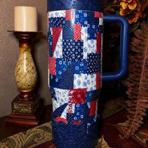 May include: A tall Stanley tumbler featuring a red, white, and blue patchwork quilt pattern. The cup has a dark blue handle, a matching dark blue lid with a paisley texture, and a dark blue base with an intricate embossed floral design.