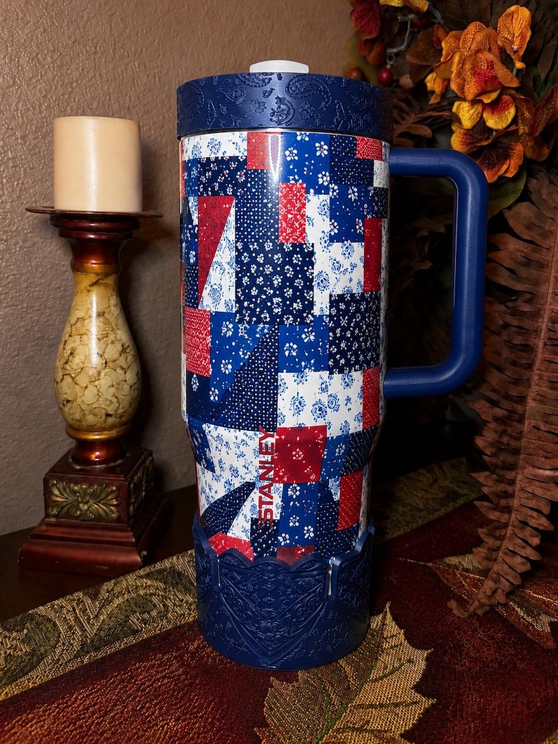 May include: A tall Stanley tumbler featuring a red, white, and blue patchwork quilt pattern. The cup has a dark blue handle, a matching dark blue lid with a paisley texture, and a dark blue base with an intricate embossed floral design.