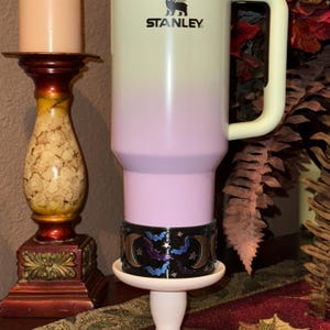 May include: A Stanley brand tumbler with a light pink and cream gradient. The tumbler has a white handle and the Stanley logo. A decorative band with moons and bats is around the base. The tumbler sits on a white stand.