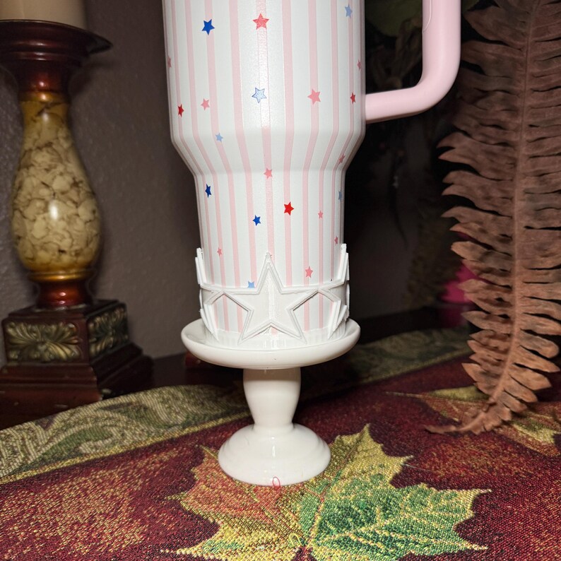 May include: A white tumbler with pink vertical stripes and red, white, and blue stars. The tumbler has a pink handle and sits on a white pedestal with a star-shaped design. The tumbler is a patriotic themed product.