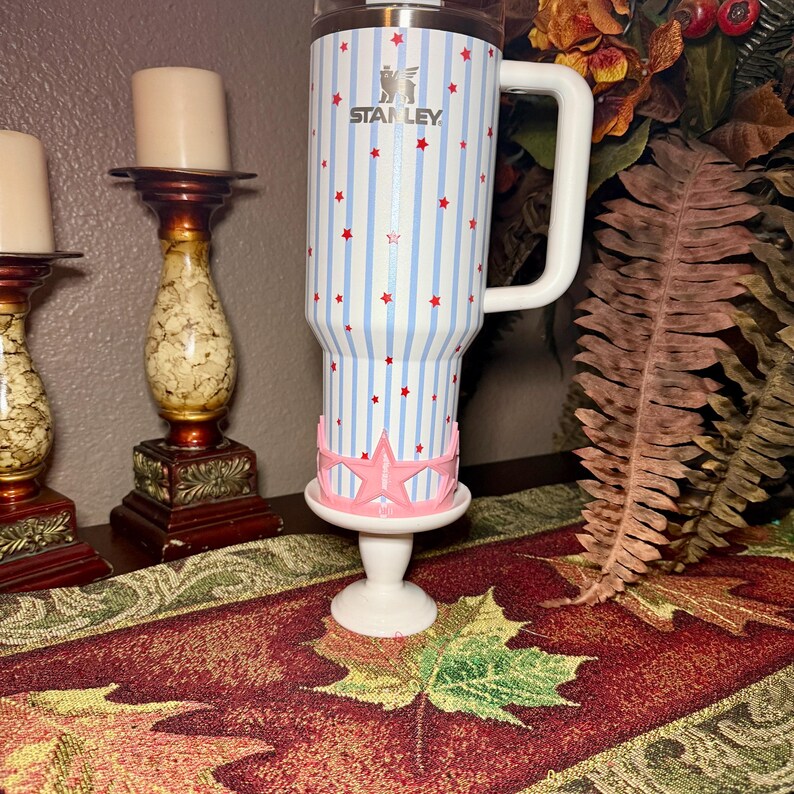 May include: A white Stanley tumbler with blue vertical stripes and red star accents. The tumbler has a white handle and is placed on a white stand with a pink star-shaped design. The Stanley logo is visible.