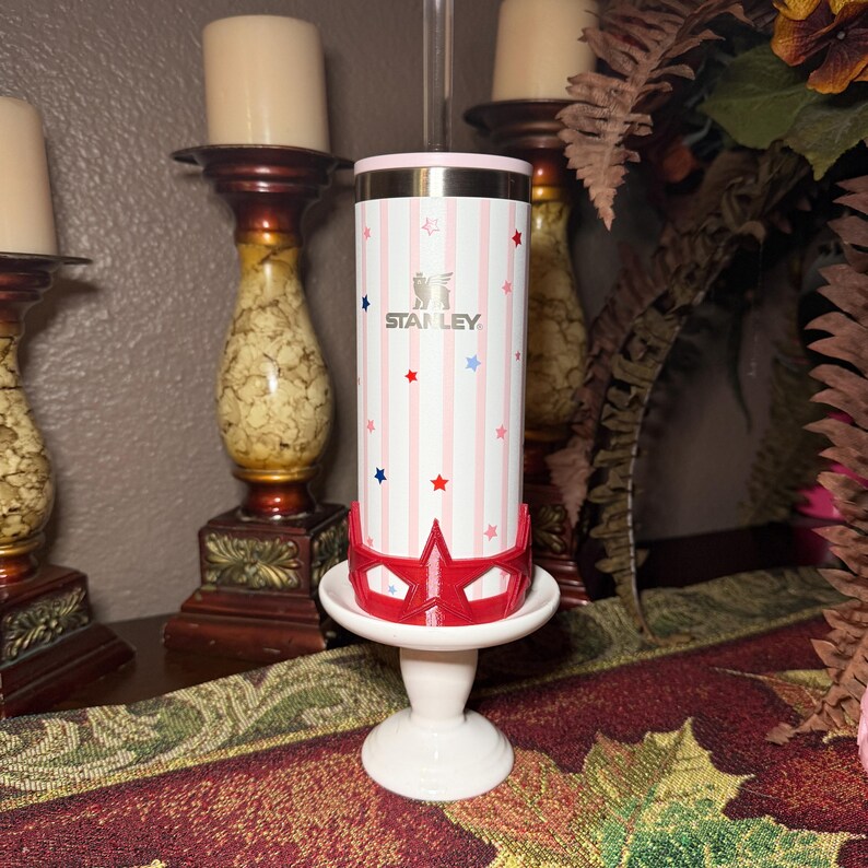 May include: A white and pink Stanley tumbler with red, white, and blue stars and stripes. The tumbler is on a white pedestal with a red star-shaped design. The Stanley logo is visible on the tumbler.