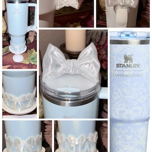 May include: A light blue Stanley Loveshackfancy tumbler with a white lace pattern. The tumbler has a white bow on the lid and a white bow-adorned base. The tumbler has a handle and a clear lid.