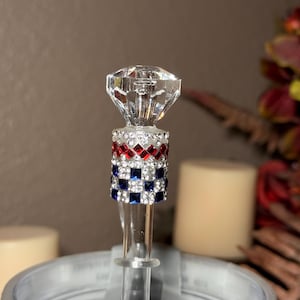 May include: A decorative bottle stopper with a clear, faceted diamond-shaped top. The stopper's stem is adorned with red, white, and blue square rhinestones. The design is reminiscent of a flag.