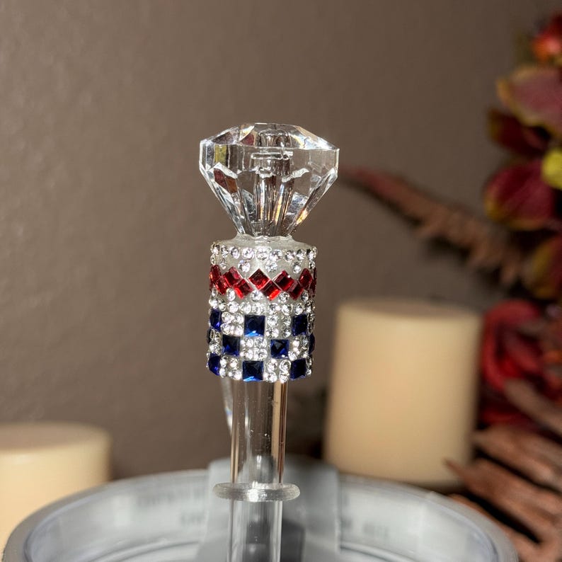 May include: A decorative bottle stopper with a clear, faceted diamond-shaped top. The stopper's stem is adorned with red, white, and blue square rhinestones. The design is reminiscent of a flag.
