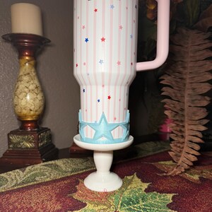 May include: A white tumbler with a pink handle, decorated with red and blue stars and pink stripes. The tumbler sits on a white pedestal with a light blue star-shaped crown.