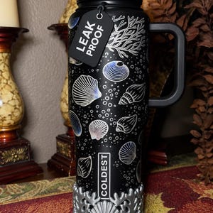 May include: A black insulated tumbler with a handle, straw, and a black tag reading LEAK PROOF. The surface features white line art of seashells, coral, and bubbles. The base has a silver-colored decorative wrap with a raised seashell and coral design.