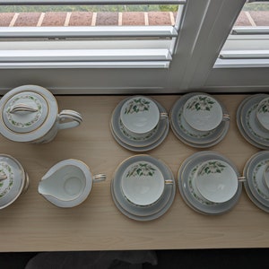 May include: A white porcelain tea set with a floral design, including a teapot, sugar bowl, creamer, and six teacups with saucers. The set features gold trim and a delicate green and white floral pattern, arranged on a light wood surface.
