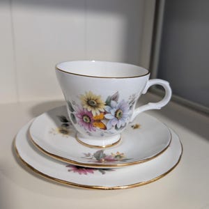 May include: A white teacup, saucer, and side plate set with a floral pattern in yellow, pink, and blue. The set has gold edging. The teacup sits on the saucer, with the side plate beneath.