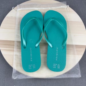 May include: A pair of teal flip-flops with a textured footbed and thin straps. The flip-flops are displayed on a light wooden surface, partially covered by a transparent drawstring bag. The brand name "PRIMARK" is printed on each flip-flop.