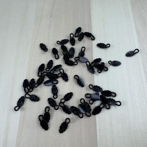 May include: A collection of small, black metal clasps scattered on a light wood surface. Each clasp has a decorative, leaf-like design and a small loop at one end. The clasps are likely used for jewelry making or other craft projects.