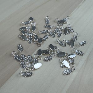 Op de afbeelding: Een verzameling kleine, zilverkleurige metalen bedels verspreid over een licht houten oppervlak. De bedels hebben een decoratief, ovaalvormig ontwerp met een lus aan één uiteinde, geschikt voor het maken van sieraden of knutselprojecten.