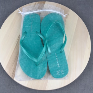 May include: Teal flip-flops in a clear mesh bag, presented on a light wood-grain surface. The flip-flops have a textured surface and a simple thong design. The brand name "Primark" is visible on one of the flip-flops.