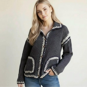 Pode incluir: Um cardigan cinza escuro com botões, acabamentos em crochê branco e bolsos. A gola e os punhos também são adornados com crochê branco. O cardigan é feito de um tecido de malha texturizado.