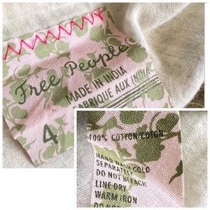 以下が含まれることがあります： 「Free People」と「Made in India」の文字が入ったピンクとグリーンの布製タグのクローズアップ。タグには花柄模様があり、ピンクの糸で縫われています。「4」の数字が見えます。