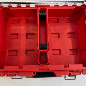 May include: A red plastic tool box with a clear lid. The interior is divided into compartments and features a black divider and several rectangular slots. The box has a metal latch and handle.