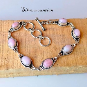May include: A silver bracelet with a toggle clasp features six oval-shaped pink opal stones set in silver bezels. The bracelet is on a wooden surface.