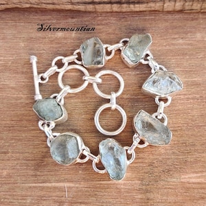 Pode incluir: Um bracelete de corrente de prata com fecho de correr apresenta pedras preciosas de água-marinha facetadas. O bracelete está sobre uma superfície de madeira.