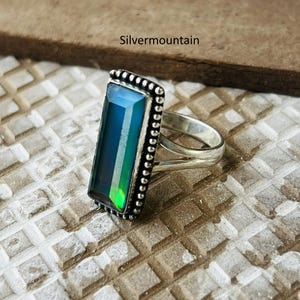 Peut inclure: Une bague en argent avec une pierre précieuse rectangulaire changeant de couleur. La pierre passe du bleu au vert. La bague a une bordure perlée et une tige fendue. Le texte "Silvermountain" est visible.