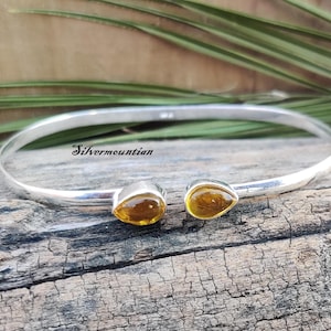 Op de afbeelding: Een zilveren manchetarmband met twee druppelvormige, amberkleurige edelstenen. De armband is open en heeft de tekst "Silvermountian". De armband wordt getoond op een verweerd houten oppervlak.