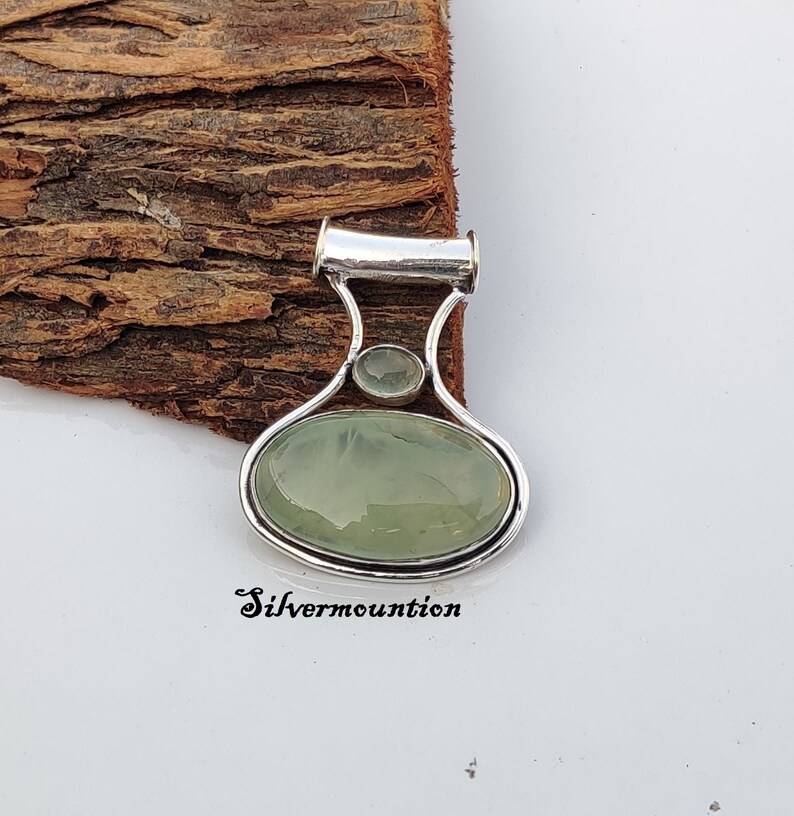 May include: A silver pendant featuring a light green oval gemstone and a smaller round gemstone. The pendant has a bail for attaching to a chain.