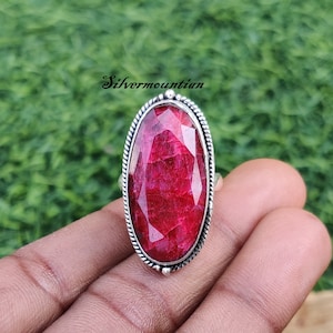 May include: A silver ring with a large, oval-shaped, red ruby gemstone. The ring has a detailed, braided band.