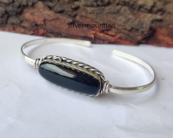 May include: A silver cuff bracelet with a large, oval, polished black stone. The stone is set in a decorative silver frame with a twisted rope design. The bracelet has a simple, open design.