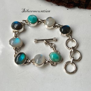 以下が含まれることがあります： 青、緑、白の色合いの楕円形の宝石が複数あしらわれた、トグルクラスプ付きのシルバーブレスレット。「Silvermountian」の文字が刻まれています。