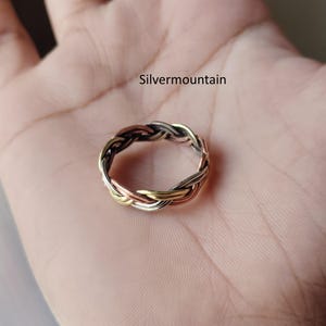 Puede incluir: Un anillo trenzado tricolor con bandas entrelazadas de metal en tonos plata, oro y cobre. El anillo presenta un patrón tejido continuo en toda su circunferencia. El texto Silvermountain es visible sobre el anillo.