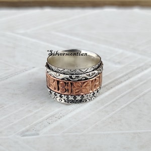 May include: A silver and copper spinning ring with intricate designs. The ring has a wide band and is made of multiple layers of metal.