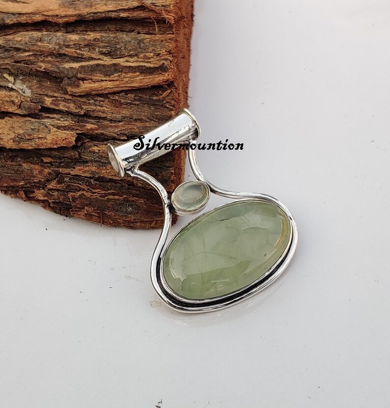 May include: A silver pendant featuring a green oval gemstone and a smaller round white gemstone. The pendant is shaped like a teardrop and has a silver bail.