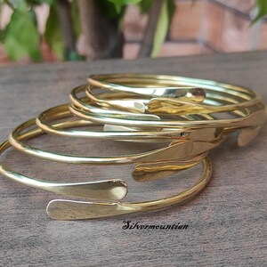 May include: A set of five gold-toned metal bangles stacked on top of each other. The bangles are all the same width and have a slightly curved shape. The bangles are smooth and shiny.