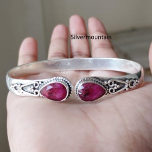 Può includere: Un bracciale rigido in argento con due pietre preziose rosa scuro sfaccettate a forma di goccia alle estremità aperte. La fascia in metallo presenta intricati dettagli decorativi in filigrana e filo intrecciato. Il testo Silvermountain è visibile.