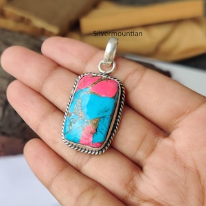May include: A rectangular pendant with a vibrant turquoise, pink, and gold stone set in a silver frame. The stone has a unique, marbled appearance. The pendant has a silver bail for attaching to a chain. The frame has a twisted rope design.