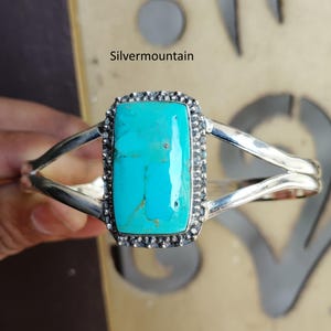 May include: A silver cuff bracelet featuring a large, rectangular turquoise stone with brown matrix veins. The stone is set in a beaded silver frame, and the band splits into two polished silver arms. The text Silvermountain is visible above.
