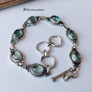 Peut inclure: Un bracelet en argent avec un fermoir à glissière présente des pierres précieuses vertes de forme ovale serties dans des chatons en argent. Le bracelet a un design à maillons de chaîne.